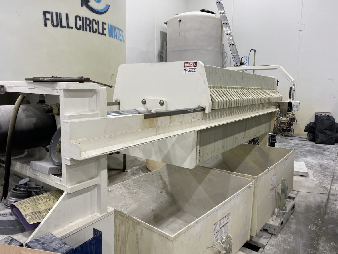 A side angle view of a filter press. Cake carts sit below it and a large white tank in the background says Full Circle Water.