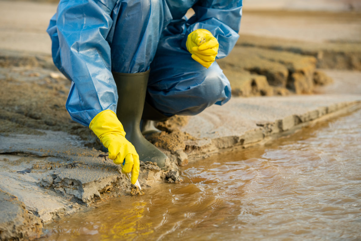 Person in a blue plastic suit with yellow gloves and rubber boots leaning over a body of dirty water